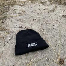 Indlæs billede til gallerivisning Sort beanie med hvidt logo på forsiden til kvinder og mænd. Huen ligger på en strand med forsiden opad. Huen passer perfekt som vinterhue, da den er dejlig varm. Den har stretch-fit, så den er super behagelig.