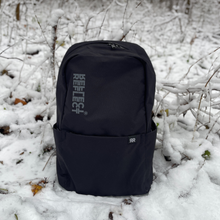 Indlæs billede til gallerivisning Miljøvenlig sort rygsæk af 100 % genanvendt plastik til kvinder og mænd. Den bæredygtige og praktiske rygsæk står oprejst i en snefyldt skov.