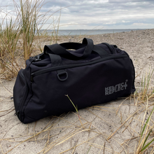 Indlæs billede til gallerivisning Sort duffel bag med gråt logo. Den ligger på en strand med havet i horisonten.