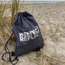 Indlæs billede til gallerivisning Miljøvenlig grå gymnastikpose af genanvendt plastik med lysegråt logo. Den står oprejst på en strand med havet i baggrunden.
